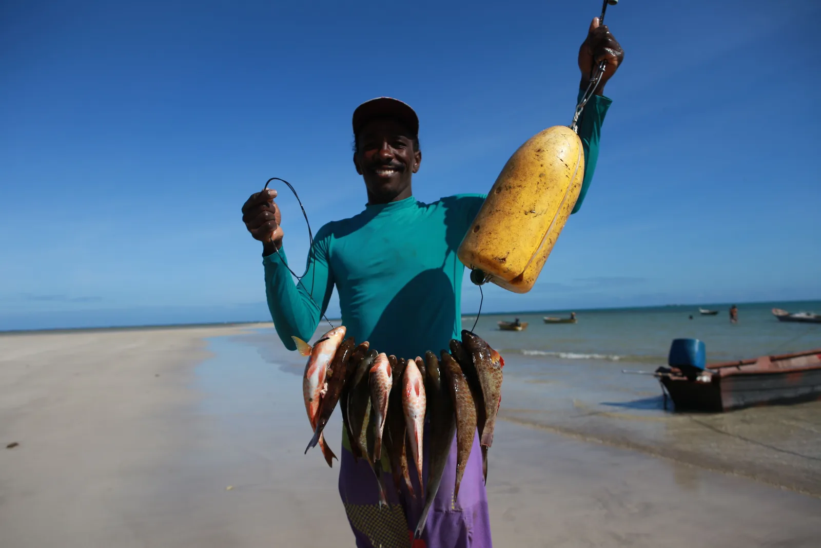 A fisherman on the sand at Garapuá holding up a line strung with the morning’s catch, a boat and a yellow float beside him
