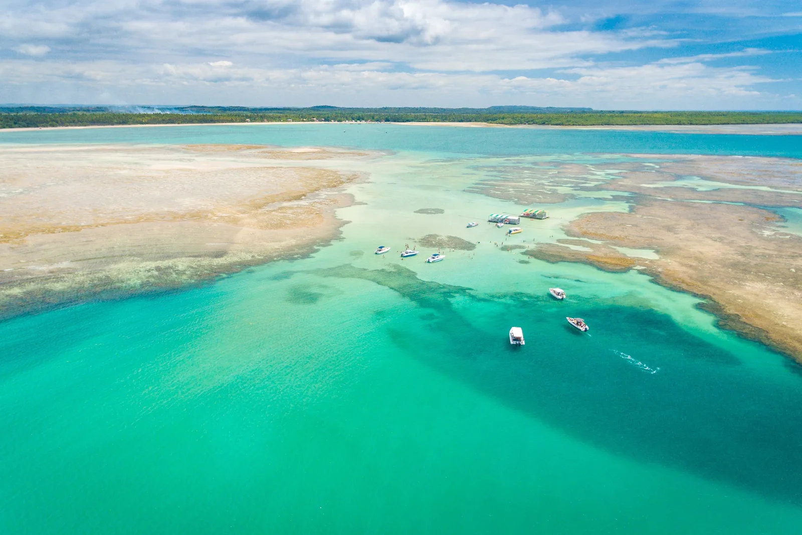 The natural pools at Garapuá from the air: a curving turquoise channel between pale sandbanks with small boats anchored on it and a speedboat leaving a wake across the shallows