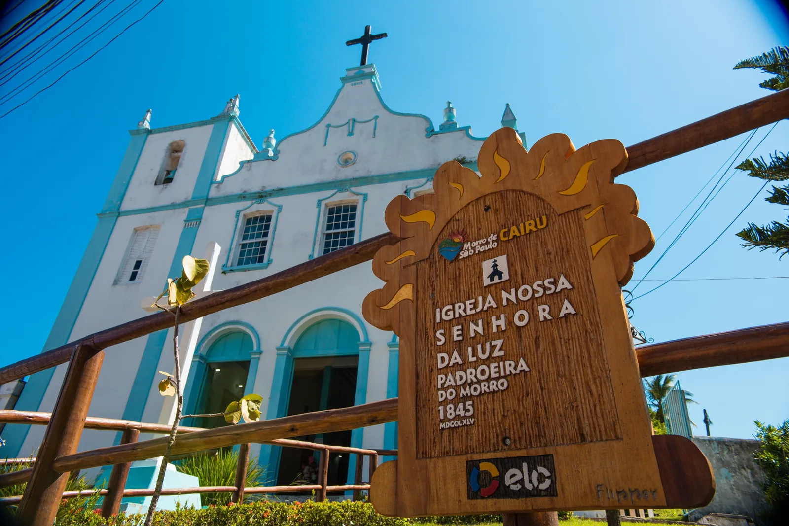 The white and pale blue facade of the parish church of Nossa Senhora da Luz in Morro de São Paulo, with a carved wooden sign in front of it naming the church and the year 1845