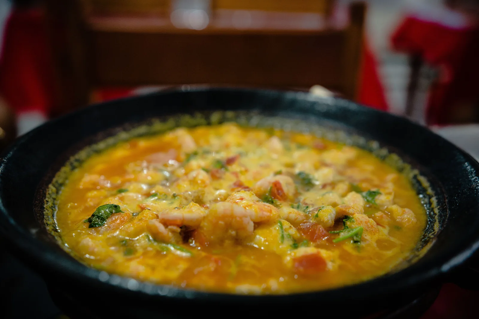 A moqueca in a black pan, the broth orange with dendê and coconut milk and full of shrimp, peppers and tomato, with a red-topped bottle behind it