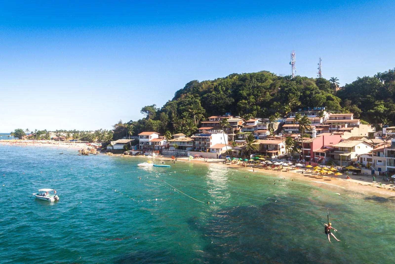 The village of Morro de São Paulo from the air above Primeira Praia: pousadas and pale buildings packed along the shore below a forested hill, with clear shallow water and a small boat offshore