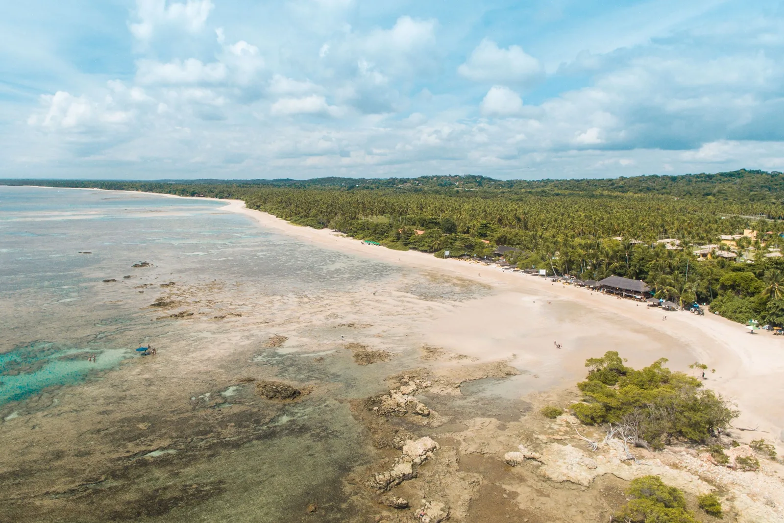 Quarta Praia from the air: a long straight beach running to the horizon with reef and pale sandbanks showing through the shallow water, and unbroken green forest behind the sand
