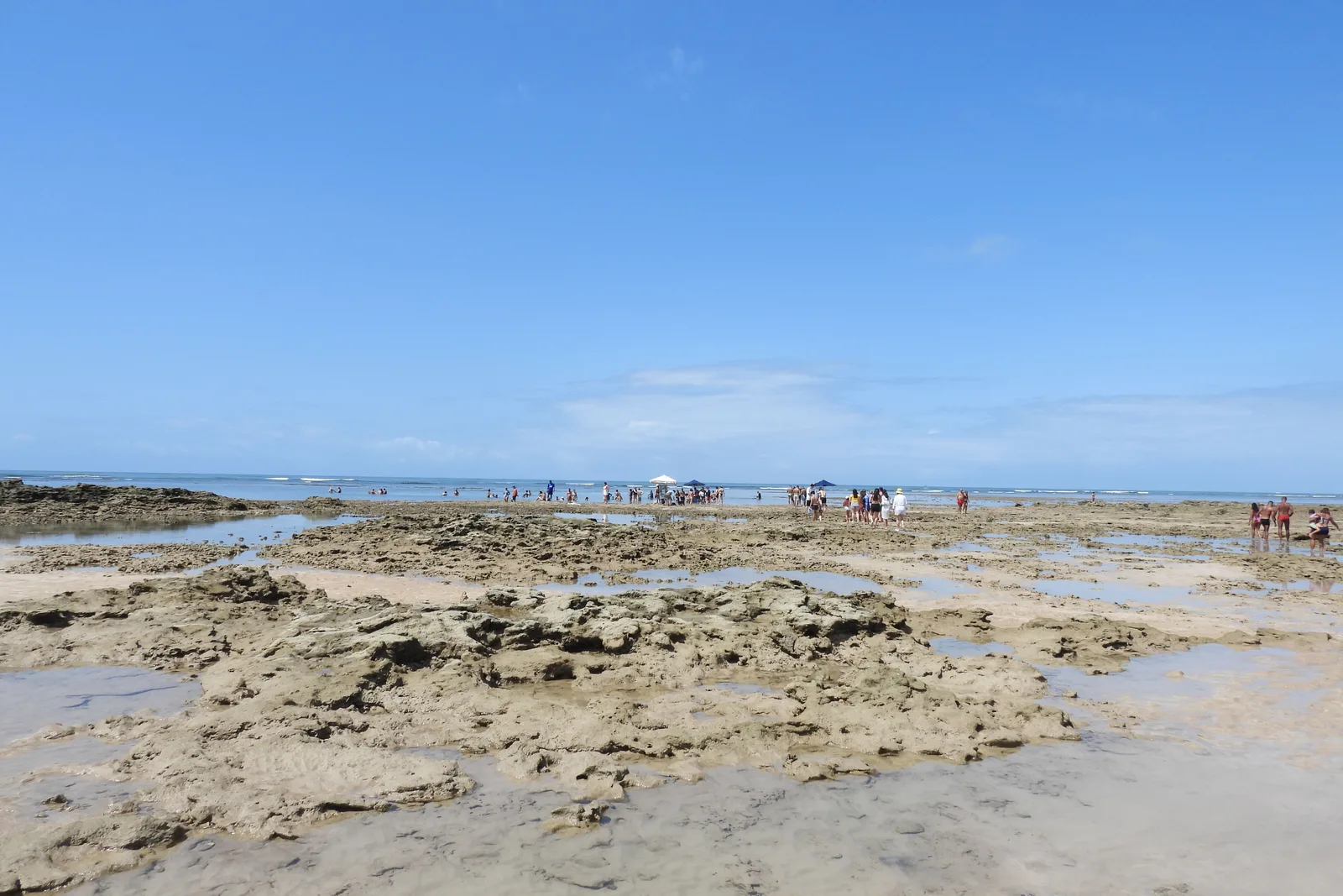 A wide shelf of reef exposed at low tide off Morro de São Paulo, people picking their way across the dark rock with shallow water standing in the hollows