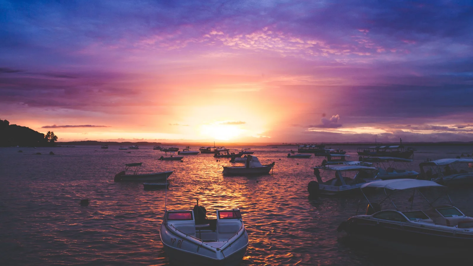 Sunset over the water at Morro de São Paulo, small boats lying at anchor in the foreground and the sun dropping behind a low line of land