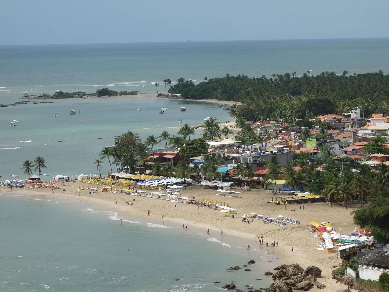 Segunda Praia from the hill above the village on a busy day: rows of colored umbrellas and loungers down the whole length of the sand, swimmers in the shallows, and the beach bars and rooftops of the village packed in behind