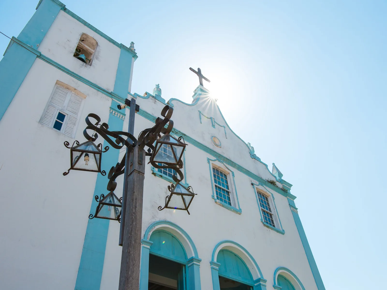 The white and blue facade of the church of Nossa Senhora da Luz in Morro de São Paulo seen from below, its bell tower and cross against a bright sky