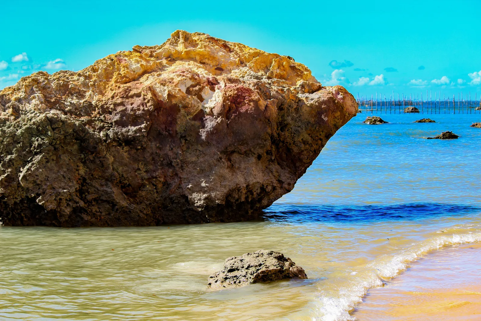 A pale rock standing on the sand at the edge of clear turquoise water at Praia da Gamboa, with oyster stakes out in the shallows behind it