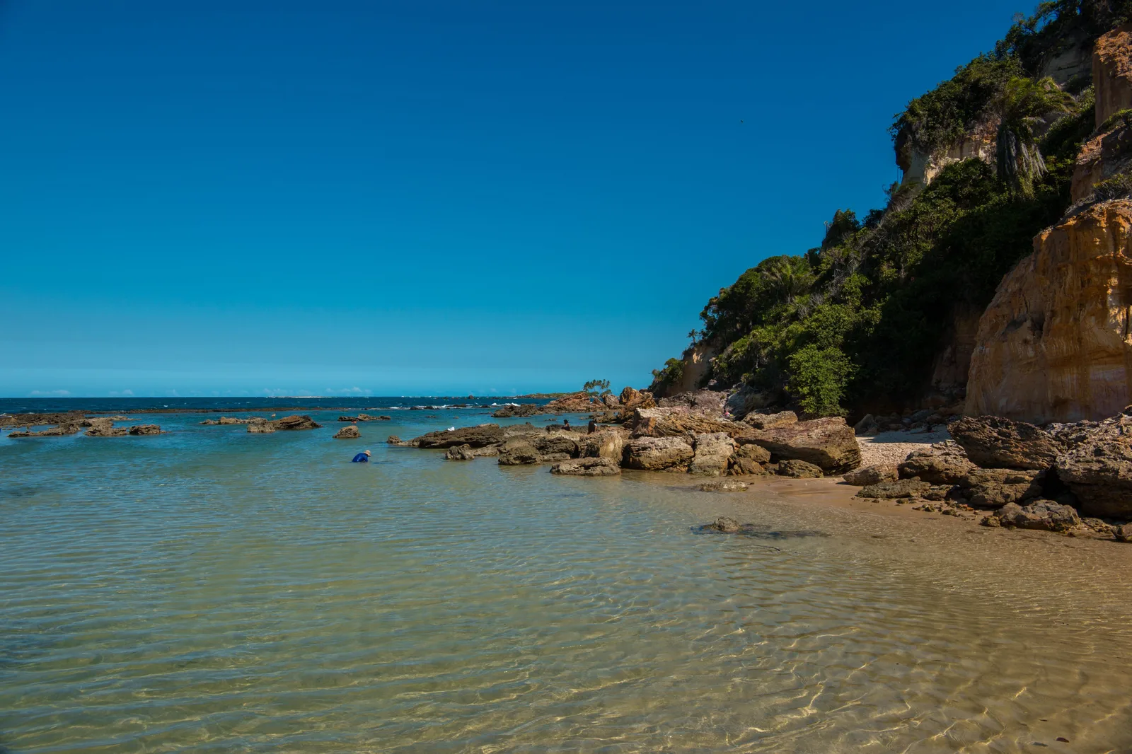 The rocky shoreline below the fort at Morro de São Paulo, clear green water over dark reef and a wooded cliff rising behind the beach