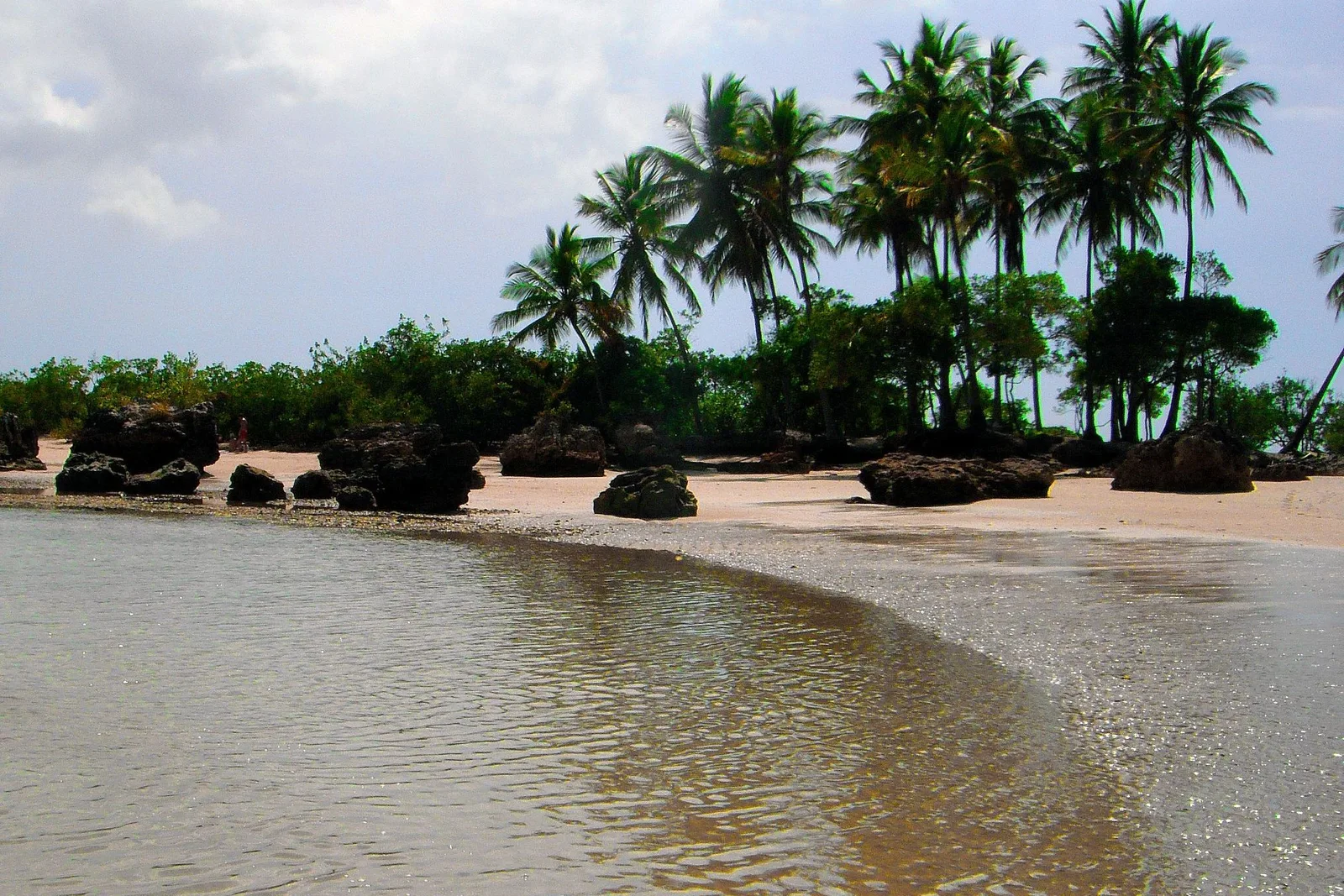 A sandbar at low tide on Segunda Praia with dark reef rock along the water’s edge and a headland of coconut palms behind