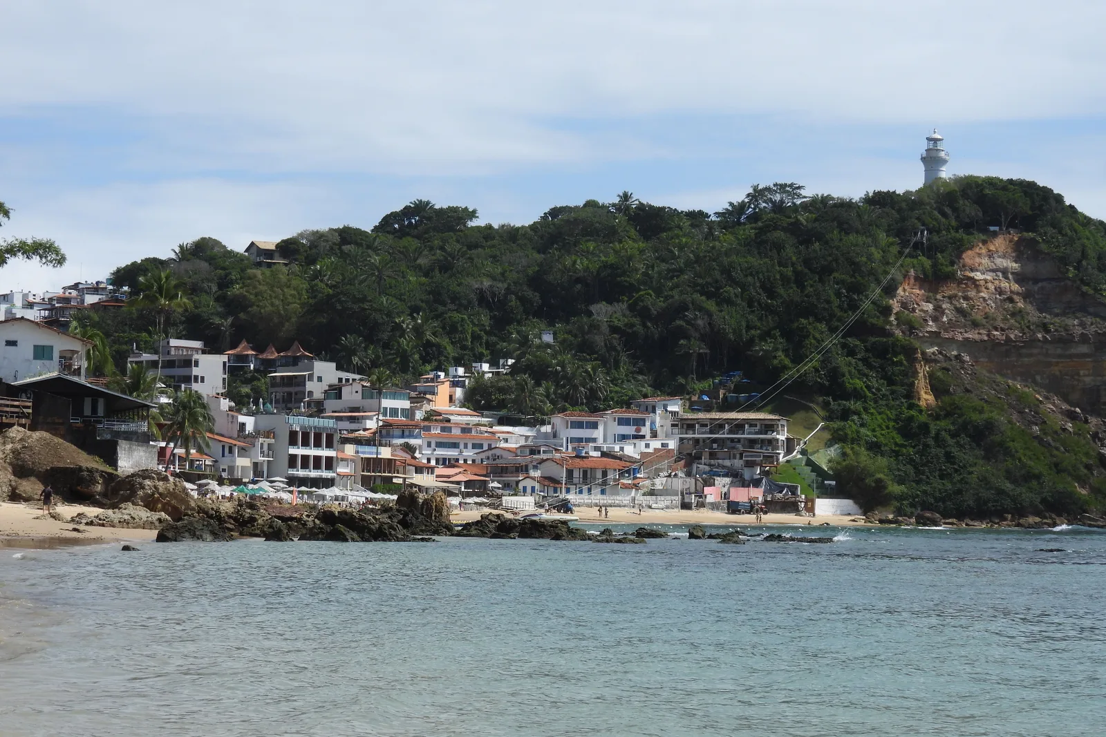 The village of Morro de São Paulo seen from the water: white and colored houses stacked up the green headland with the lighthouse on the ridge above them