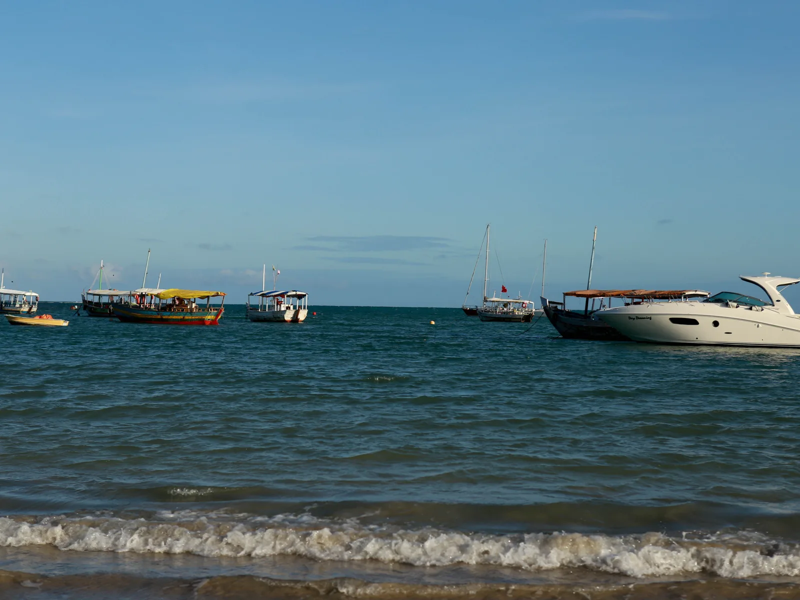 Boats lying at anchor off a beach in the Tinharé archipelago, low waves running up the sand in the foreground and a wooded shore behind them