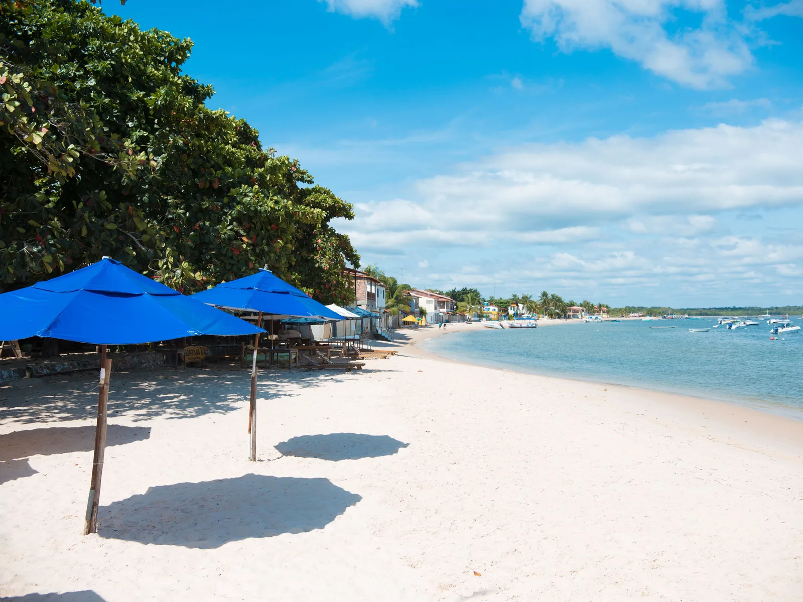 The beach at Gamboa on a bright day, blue parasols set out on white sand, small boats at anchor in flat water and a line of low buildings along the shore