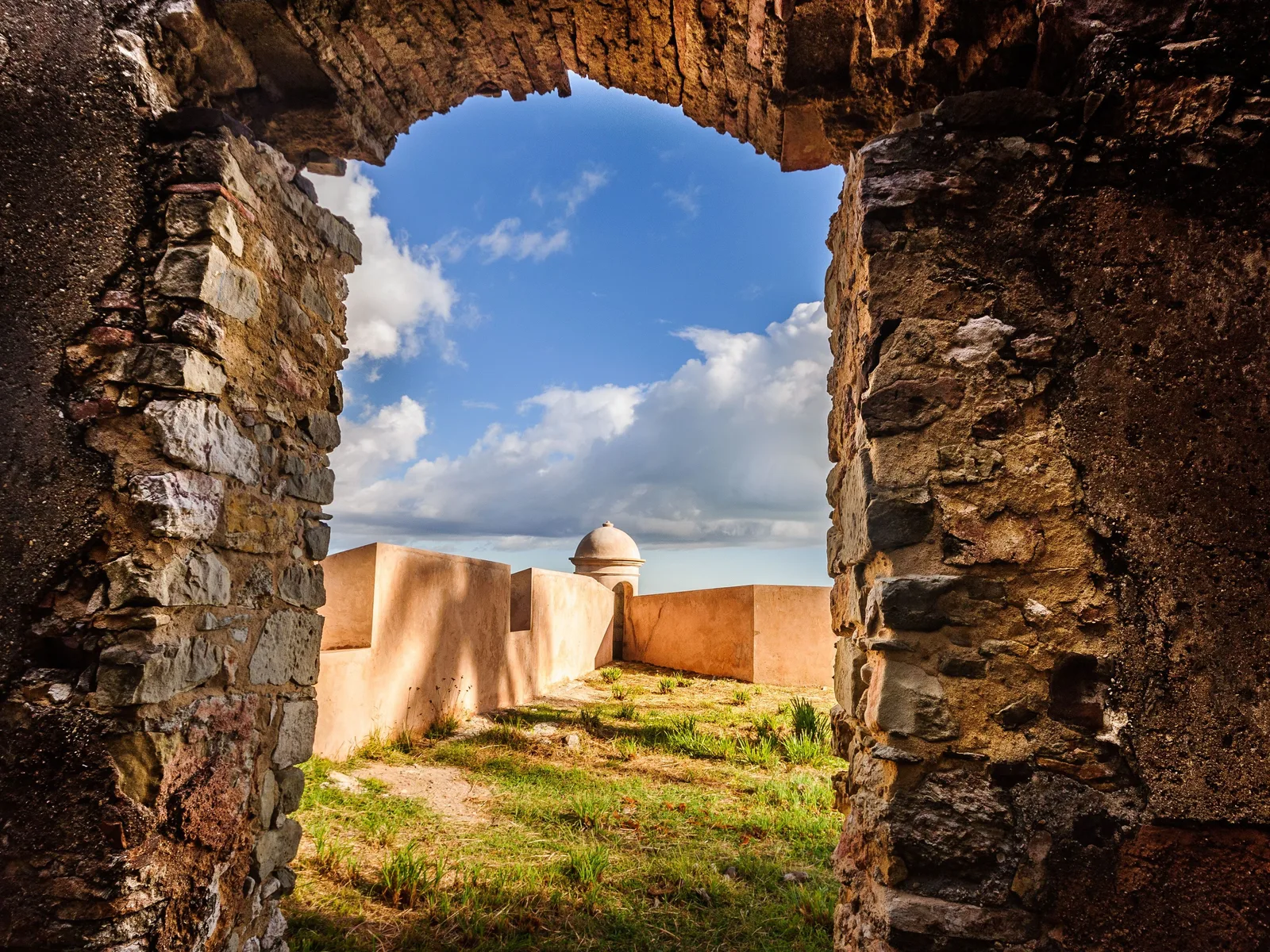 The ruined stone walls of the Fortaleza de Tapirandu at Morro de São Paulo framed through a broken archway, with two round sentry boxes standing on the rampart against a bright sky
