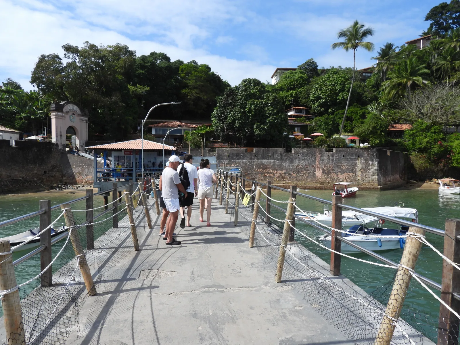 Arriving passengers walking up the causeway from the pier at Morro de São Paulo toward the arched stone gateway in the old fortress wall, speedboats moored along the rocks either side