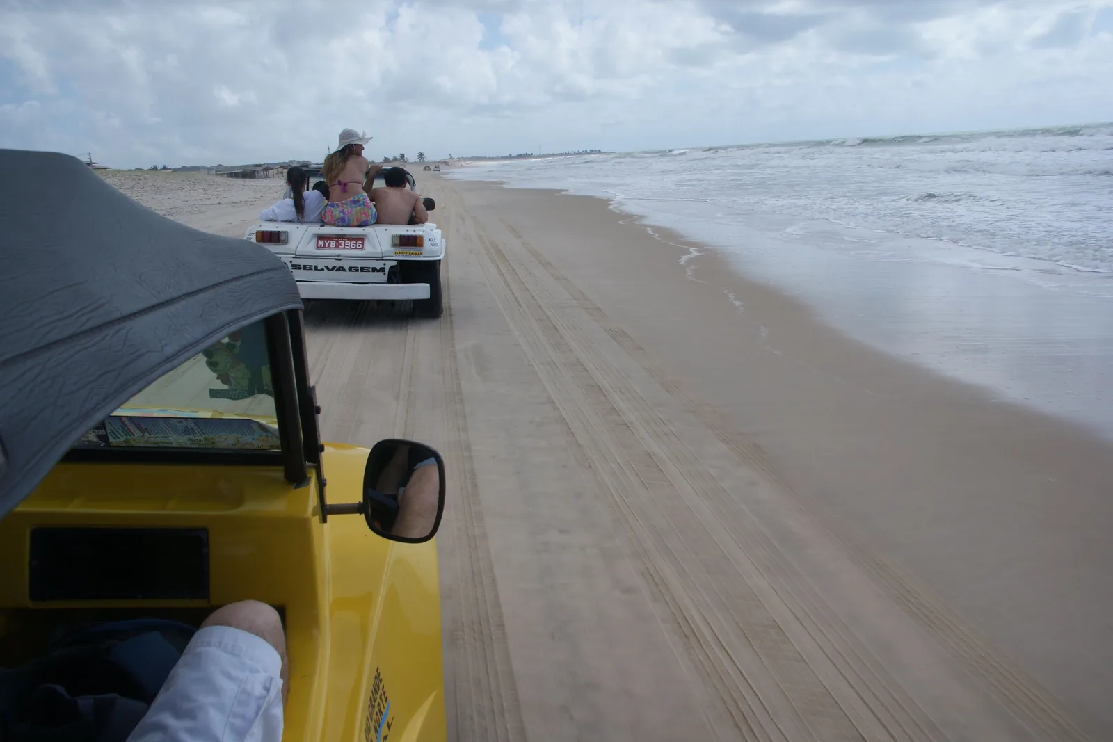 View forward from inside a yellow beach buggy with its roof up, following another buggy along a wide flat beach beside the surf
