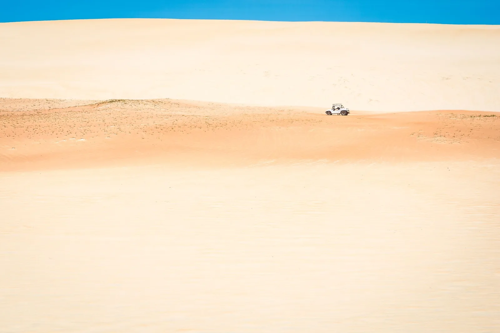 A single buggy crossing an enormous unbroken sweep of pale sand under a thin band of blue sky, small enough in the frame to show the size of the dune