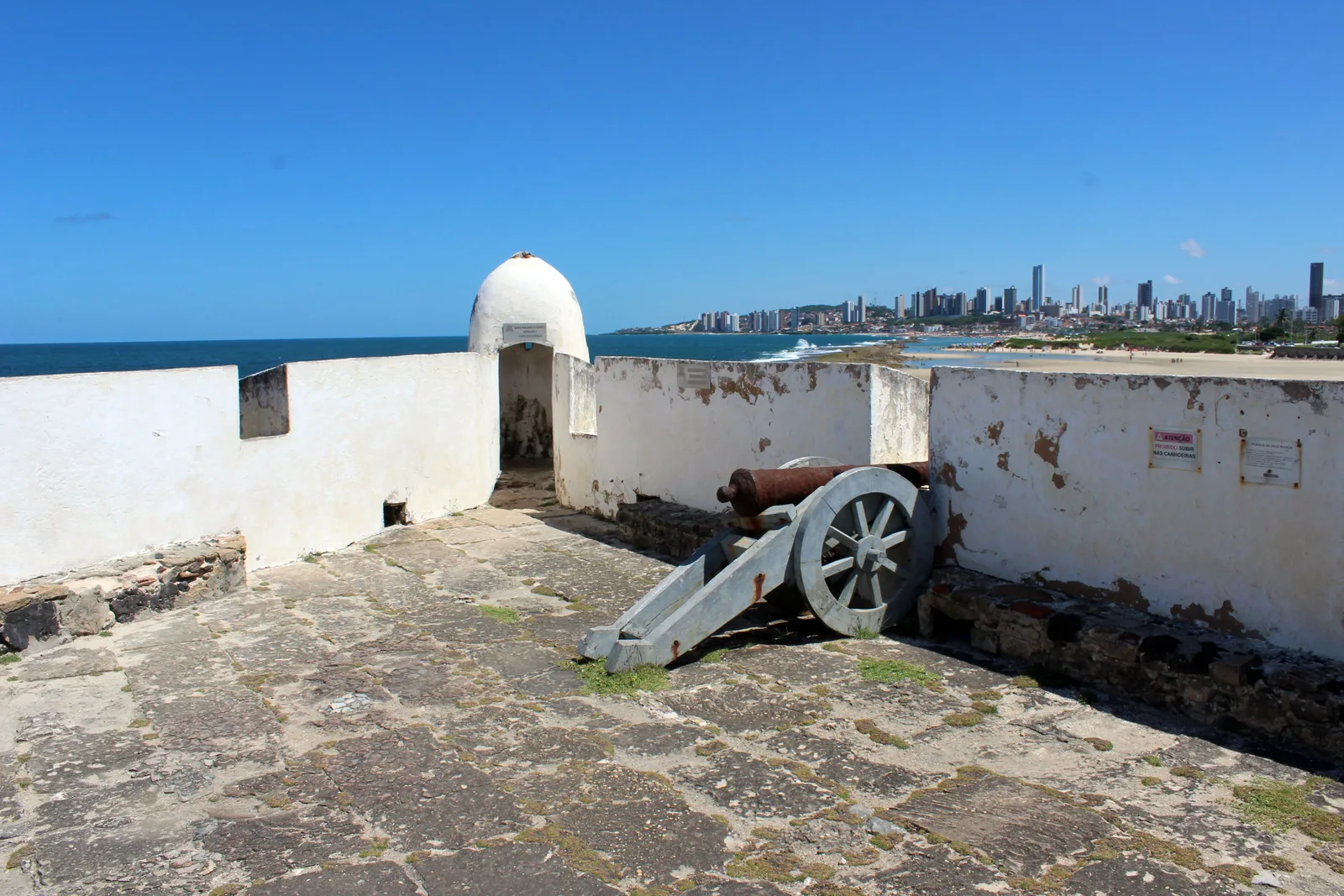 A rusted cannon on a gray spoked-wheel carriage standing on the open upper deck of the Fortaleza dos Reis Magos, with a white sentry turret behind it and the towers of Natal across the water