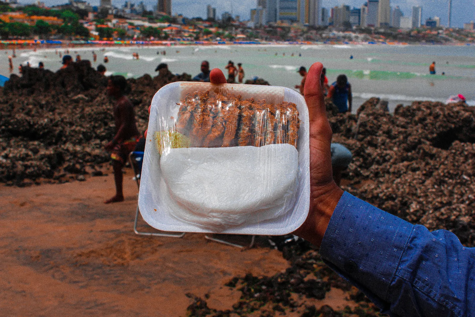 A hand holding a plastic-wrapped tray with a row of little fried fish above a folded white tapioca, on a beach of dark reef rock with bathers and the city skyline behind
