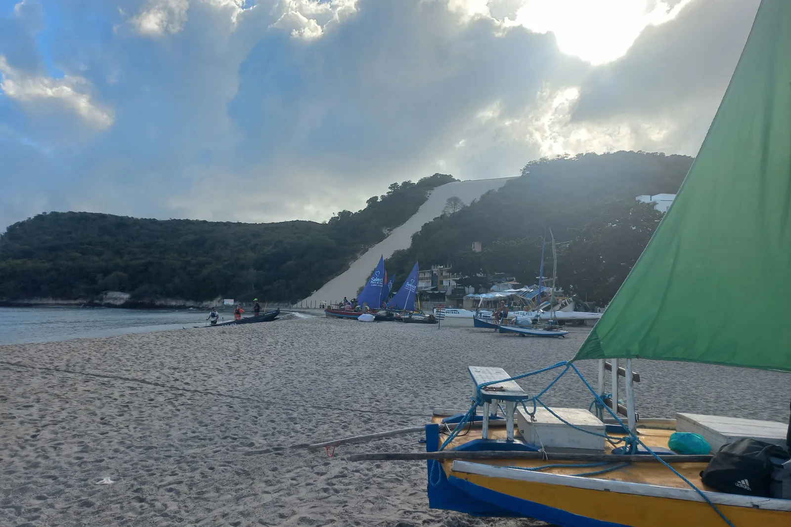 A wooden sailing boat with a green sail pulled up on the sand at Ponta Negra in the early morning, backlit, with Morro do Careca in haze behind it