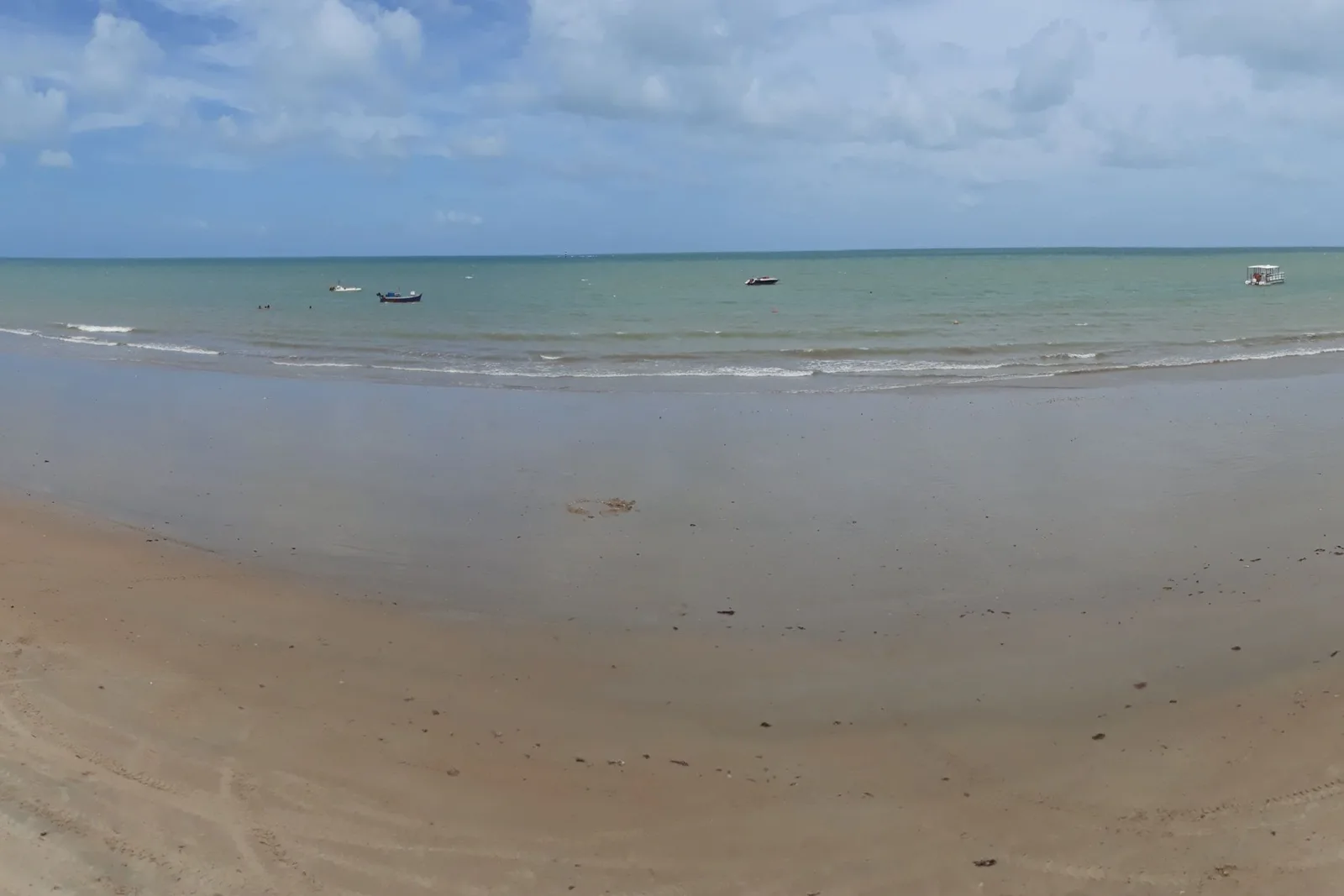 The beach at Maracajaú at low tide: a broad expanse of wet flat sand and shallow water with small boats moored far out toward the horizon