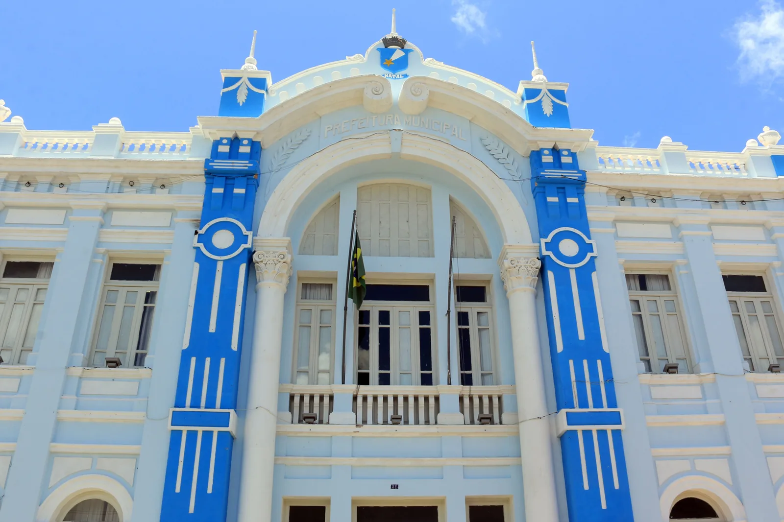 The facade of the Palácio Felipe Camarão in Natal: a civic building painted pale blue with bright blue pilasters, a tall arched central bay over a balcony and flag, shuttered windows and small pinnacles along the roofline