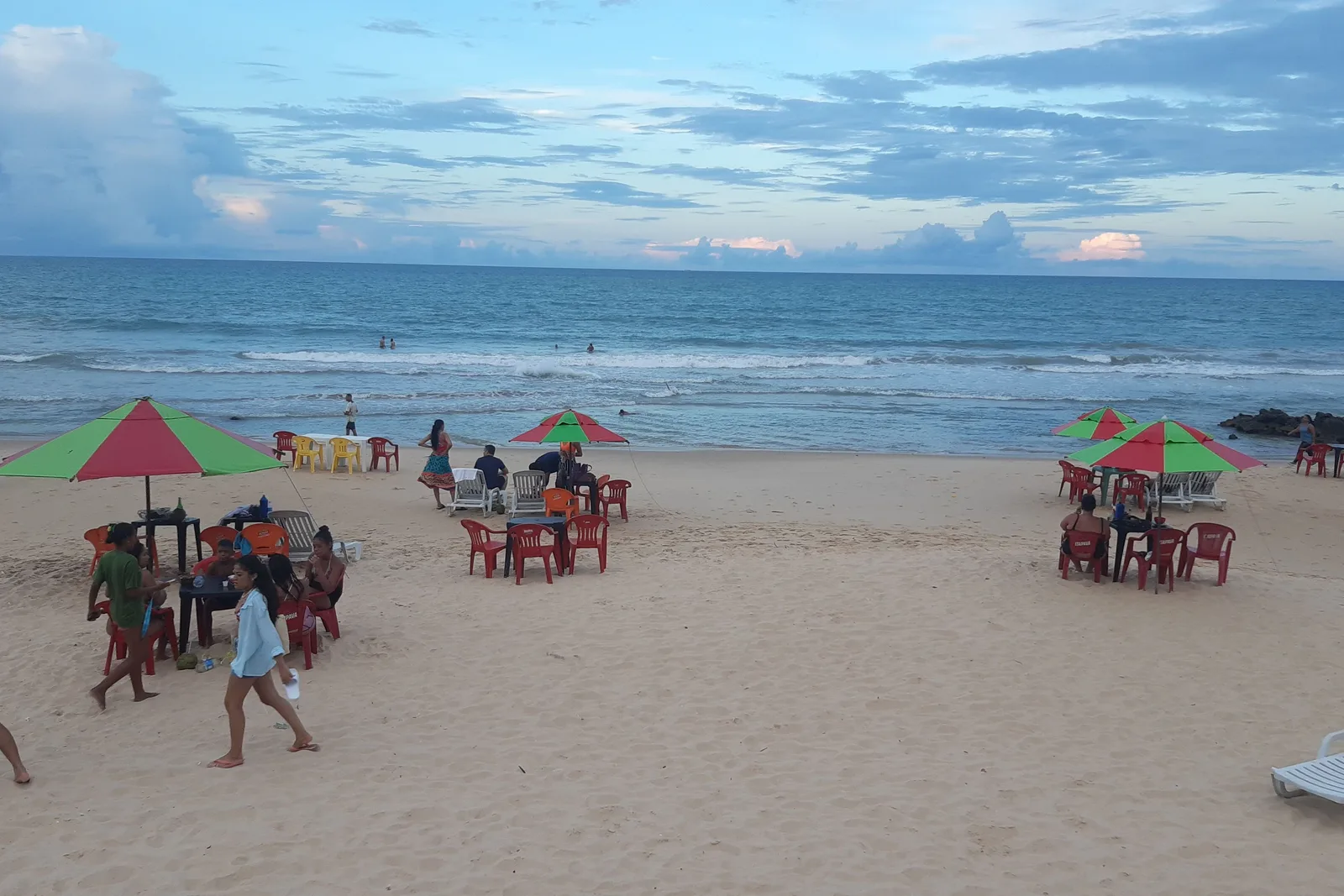Red plastic tables and chairs under red and green umbrellas on the sand at Praia do Meio in Natal, with people walking at the water’s edge and dark rocks at the far end of the beach