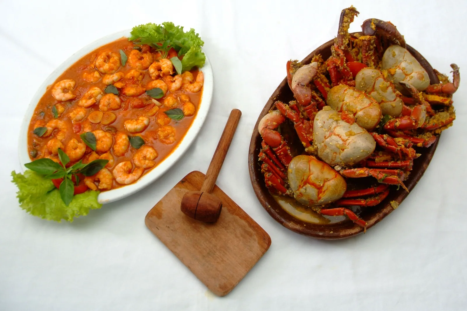 A white oval dish of shrimp in an orange sauce beside an oval clay dish of whole cooked crabs, with a small wooden mallet on a board between them