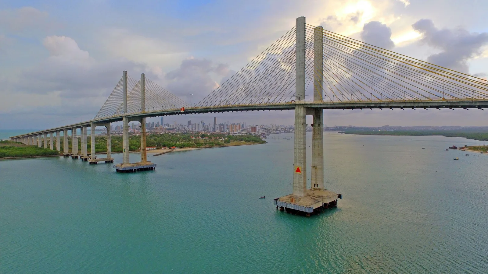The cable-stayed Newton Navarro bridge crossing the wide green mouth of the Potengi river in Natal, with its concrete piers standing in the water and the far bank beyond