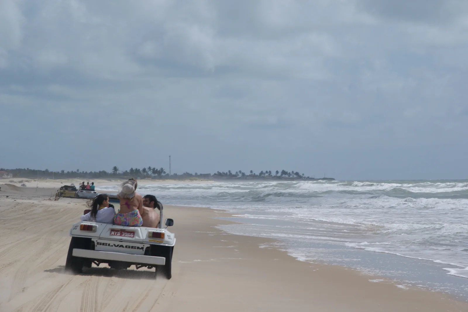 A white beach buggy carrying passengers driving along the wet sand at the edge of the surf, with a low hazy shoreline in the distance