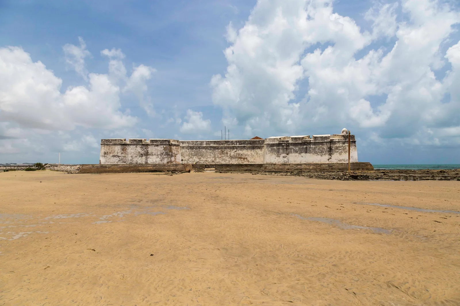 The Fortaleza dos Reis Magos seen across a vast expanse of bare wet sand exposed by a low tide, its white walls low against a cloudy sky