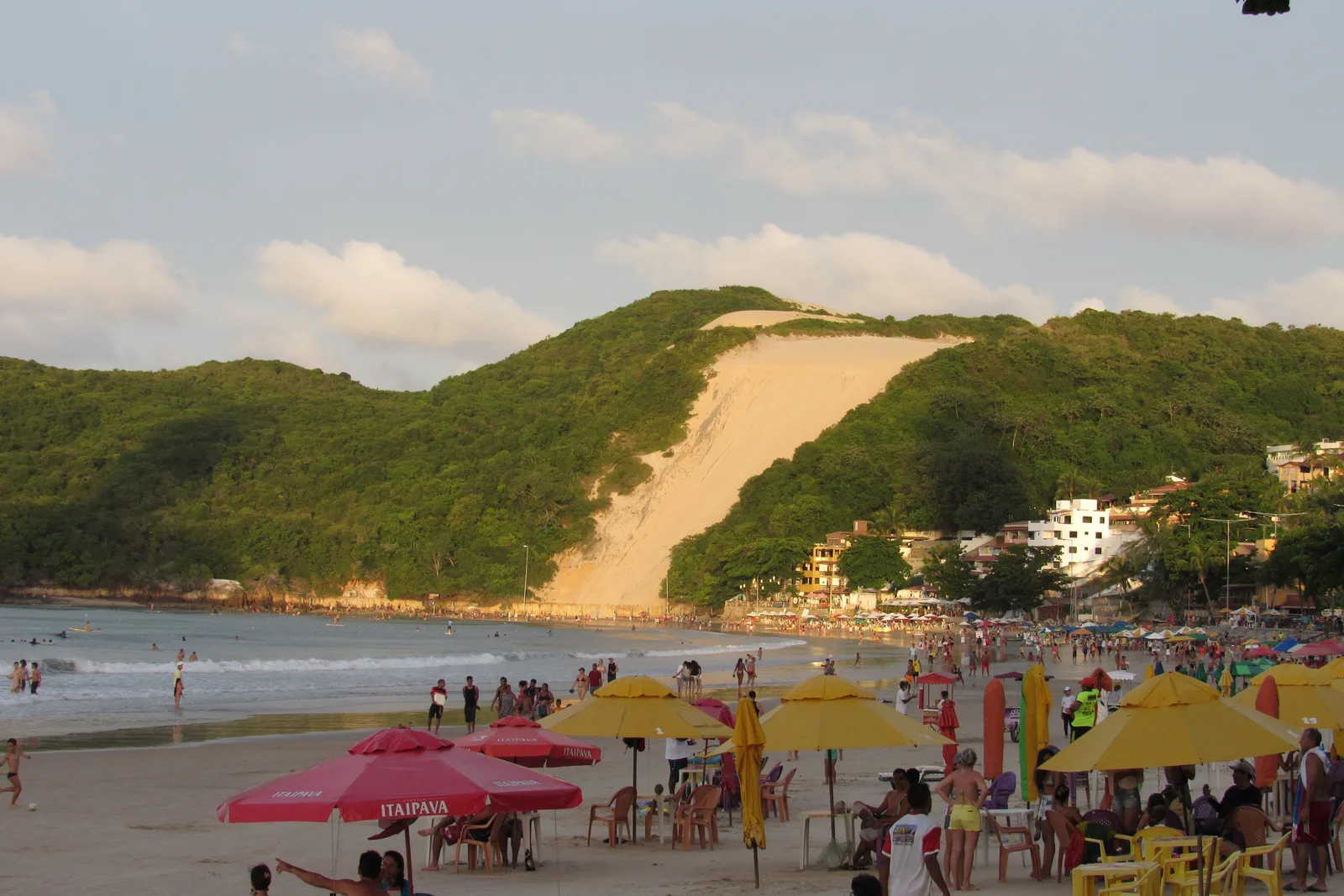 Rows of yellow and red beach umbrellas and tables on the sand at Ponta Negra, with the green and sand slope of Morro do Careca rising behind them