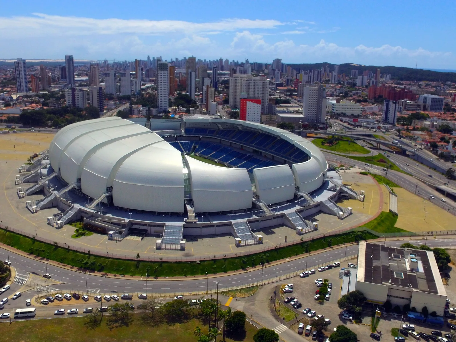 Aerial view of the Arena das Dunas stadium in Natal, a white segmented roof structure surrounded by parking lots and the city’s apartment blocks