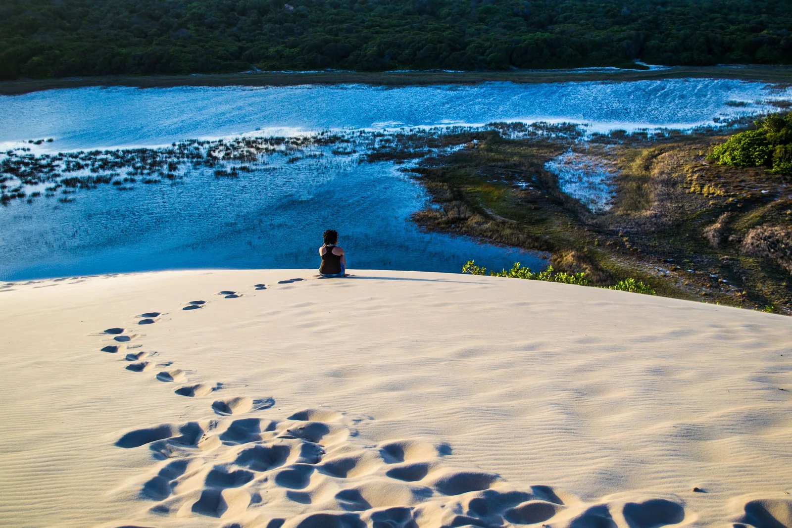 A line of footprints crossing white sand at Jenipabu toward a person sitting on the dune crest above a blue lagoon, with a forested far shore