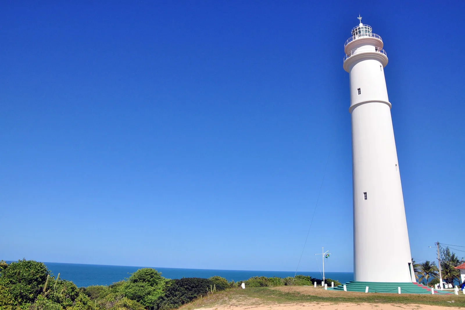 The white tower of the Farol de Mãe Luíza lighthouse against a clear blue sky, with green scrub and the sea at its foot