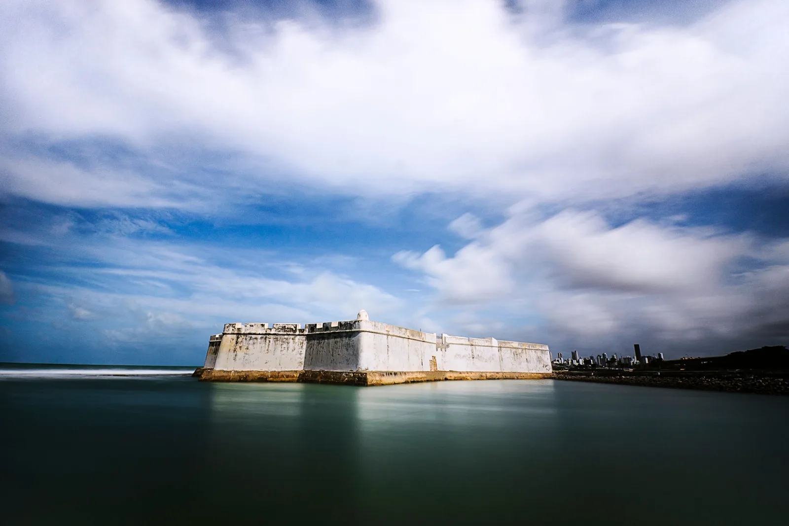 The low white walls of the Fortaleza dos Reis Magos seen across calm water under a bank of cloud, with the Natal skyline to the right
