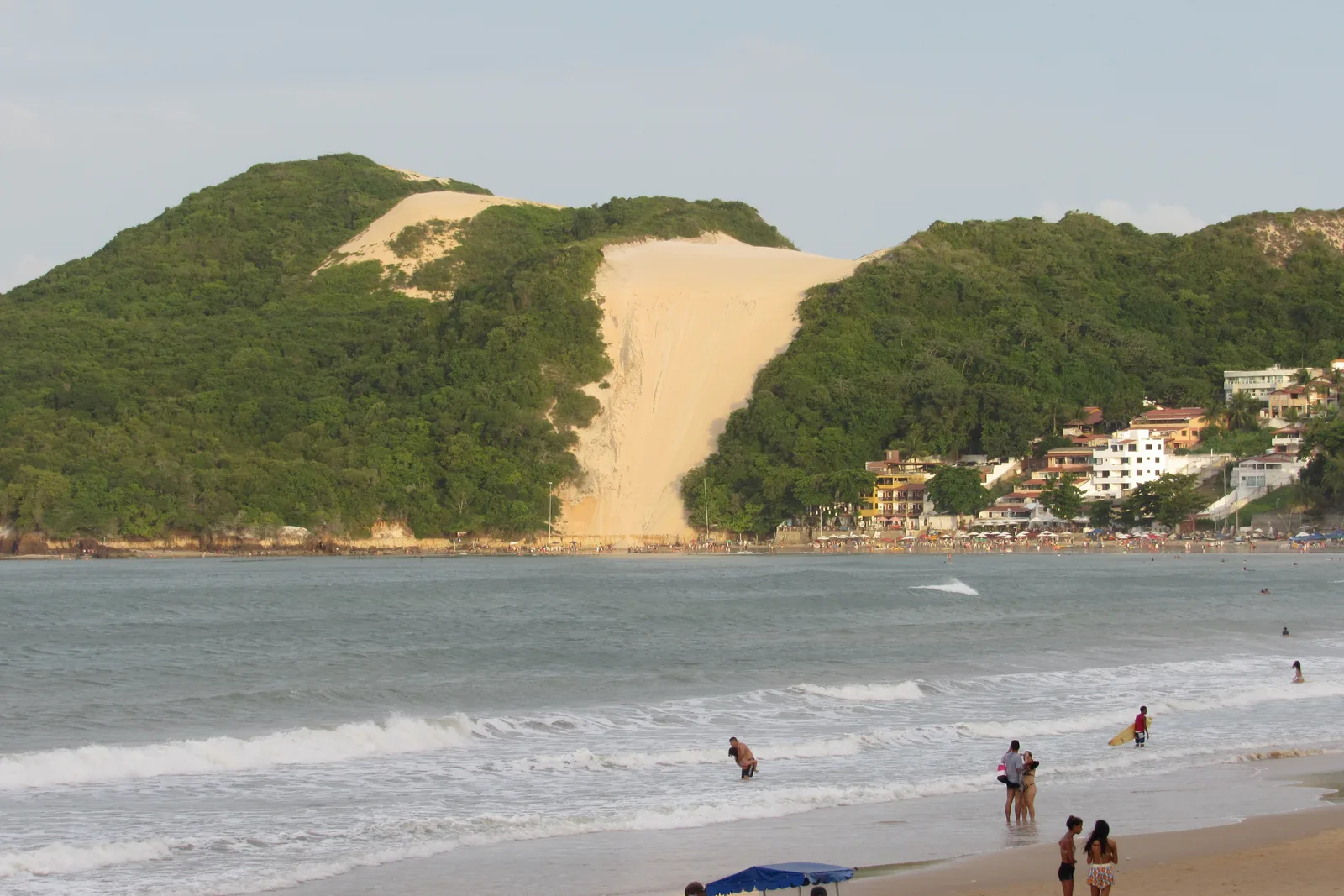 Morro do Careca from the beach at Ponta Negra: a green hill split by a bare slope of pale sand, with people standing in the shallows below