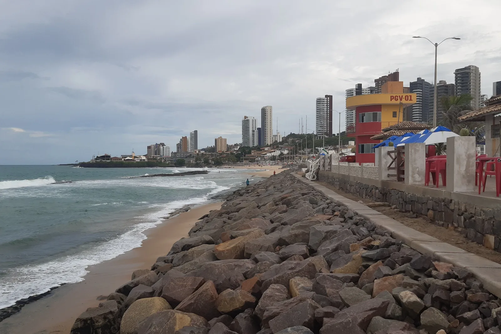 A rock sea wall running along the sand at Praia do Meio in Natal, with surf breaking beside it, a red and yellow lifeguard tower, kiosk tables and the city’s apartment towers behind