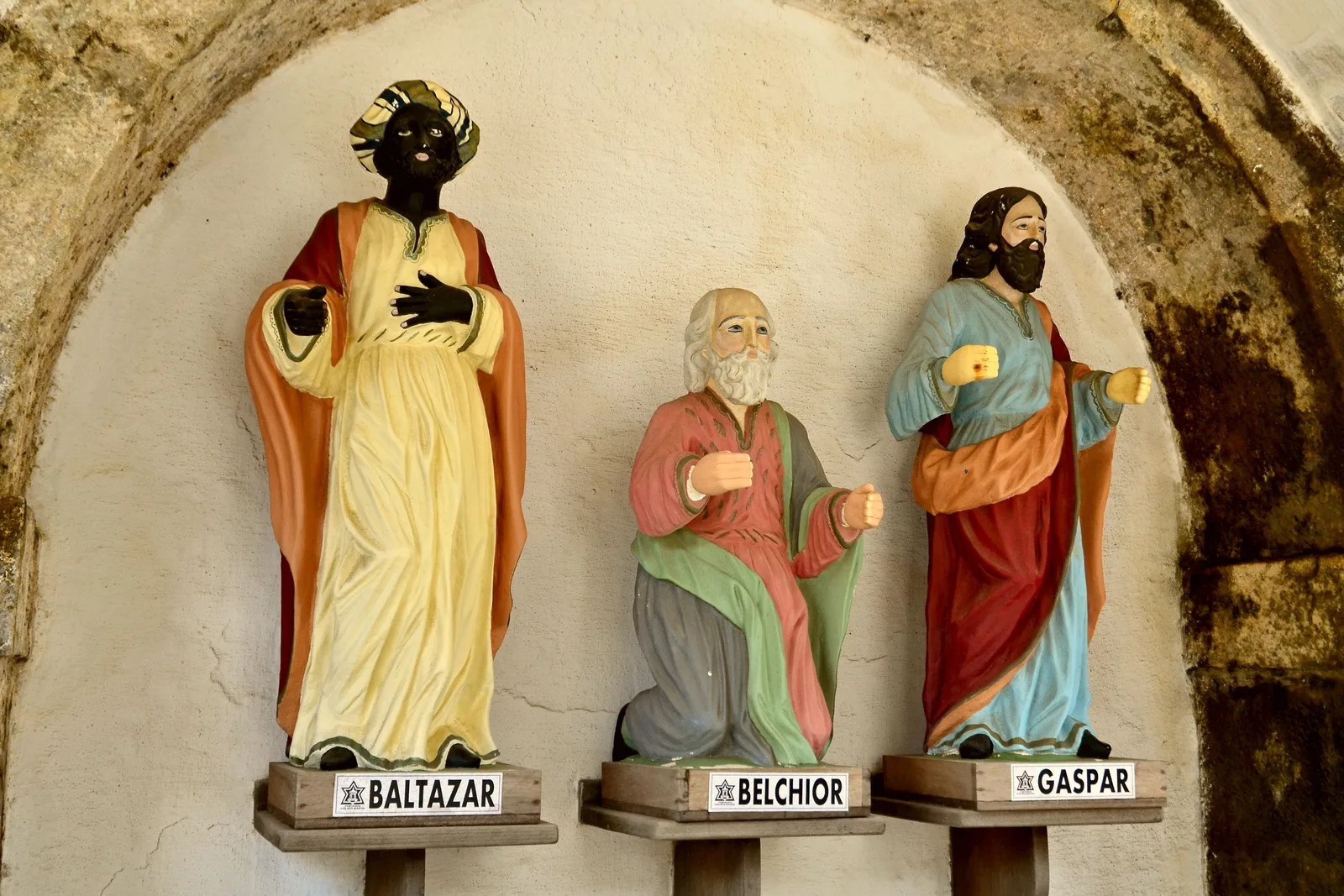 Painted statues of the three Magi on labeled pedestals, Baltazar and Gaspar standing and Belchior kneeling between them, in an arched stone niche inside the Fortaleza dos Reis Magos