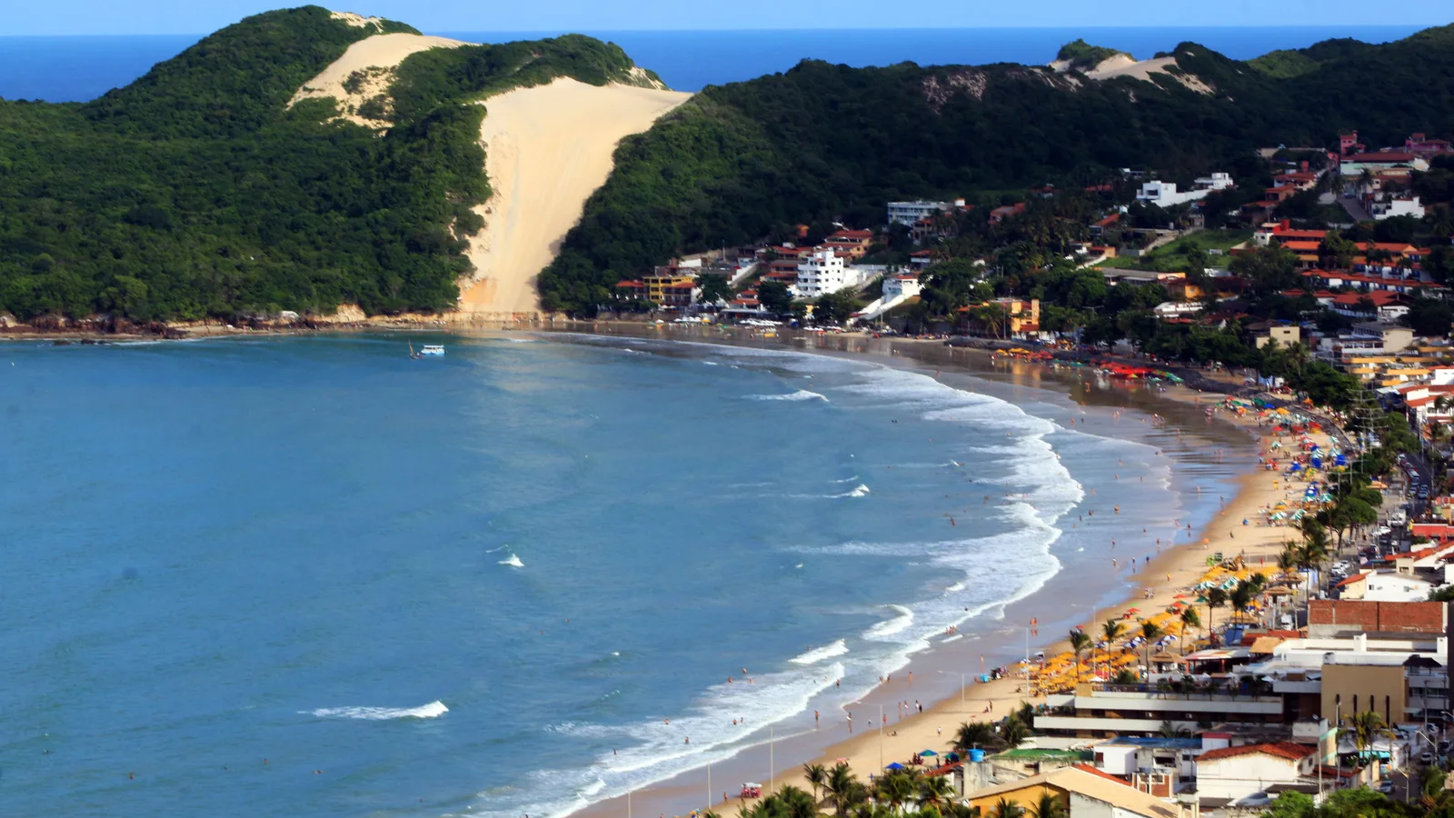 Ponta Negra in Natal from above: a curved beach lined with umbrellas and low red-roofed buildings, with the green hill of Morro do Careca and its bare stripe of sand closing the far end, and blue water breaking along the shore