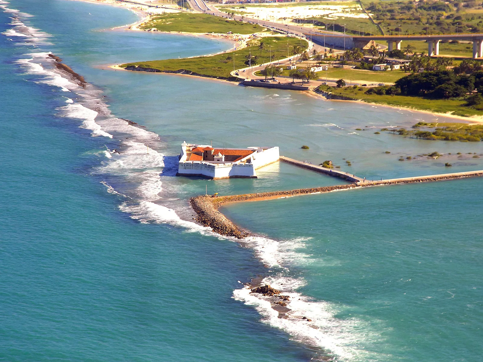 Aerial view of the Fortaleza dos Reis Magos: a low white fort with red-tiled roofs inside its walls, standing in green water at the river mouth and joined to the shore by a long walkway, with surf breaking on the reef beside it and the Newton Navarro bridge behind