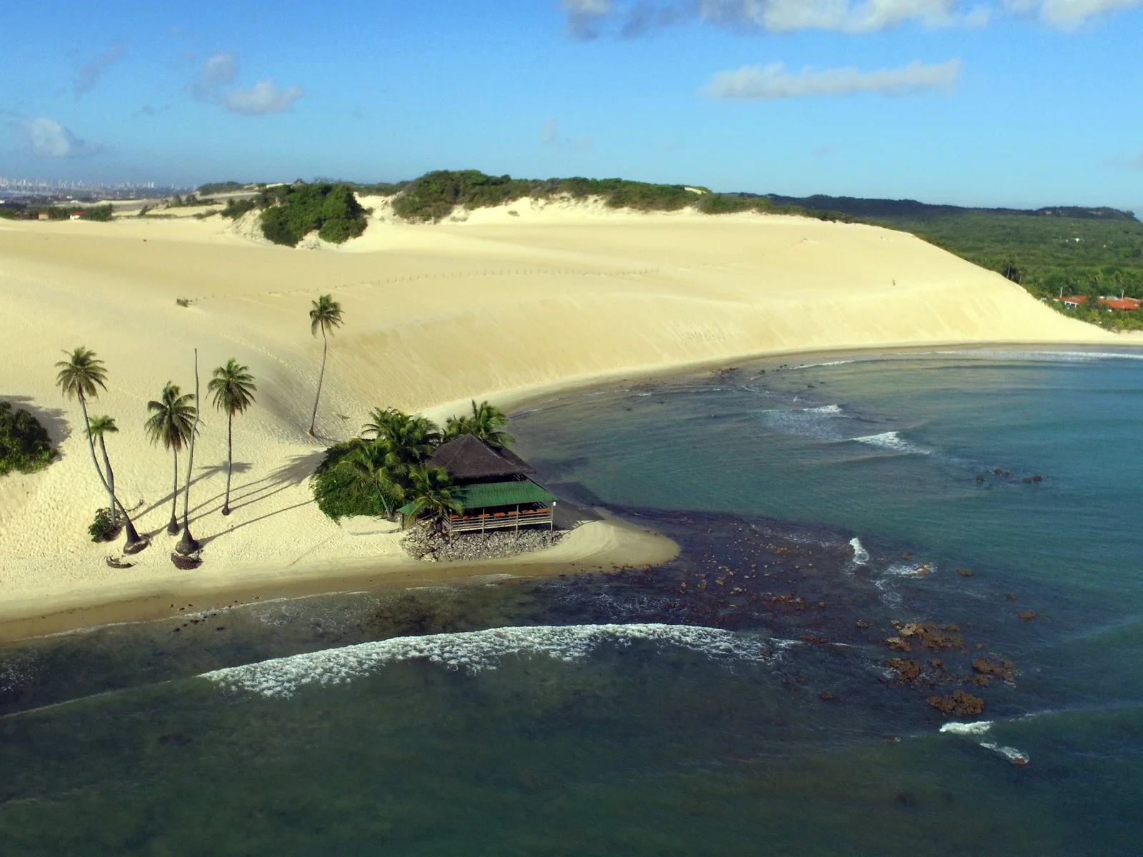 Aerial view of the Genipabu dunes: broad pale sand ridges running down to a fringe of coconut palms, a thatched shelter and shallow green water broken by dark reef