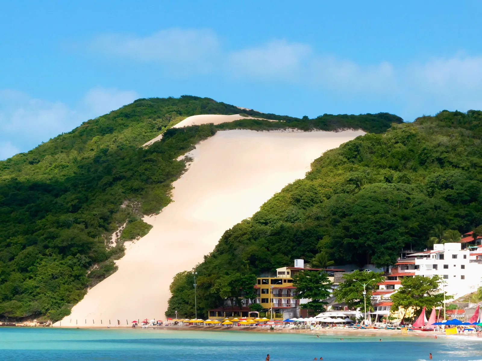 Morro do Careca at the end of Ponta Negra beach: a rounded hill covered in green scrub with a wide bare slope of pale sand running down its face to the buildings and turquoise water at its foot
