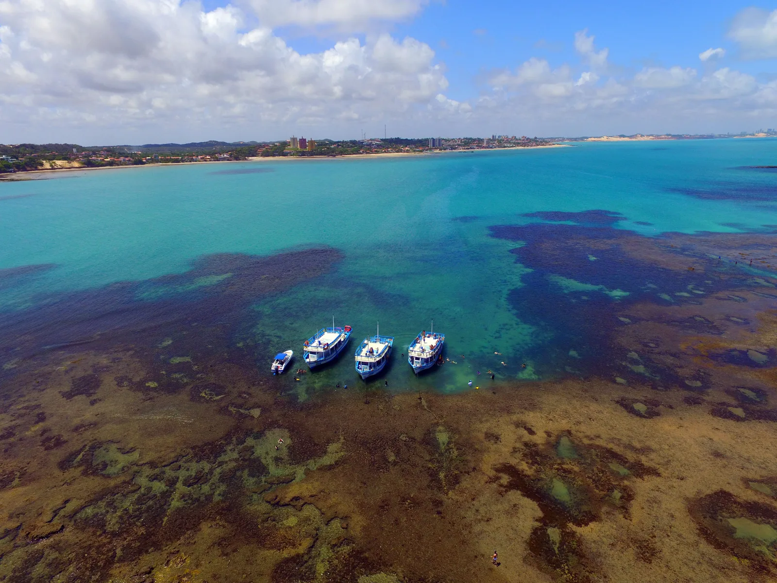 Aerial view of the parrachos off Pirangi: turquoise shallow water over dark patches of reef, with three double-deck tour boats and a small speedboat moored at the reef edge and the beach and low buildings behind