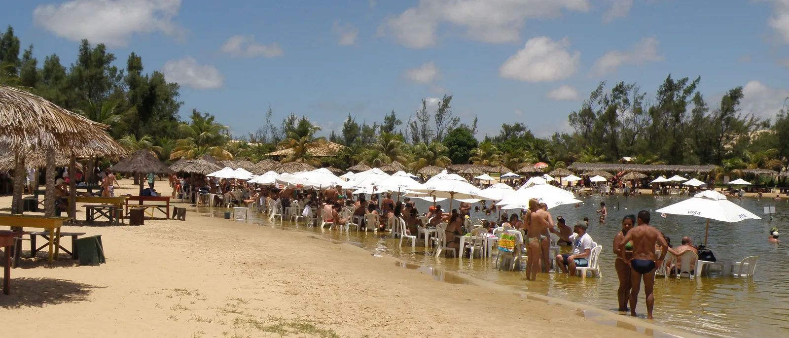 White parasols and plastic tables set out in the shallow water of a lagoon in the Genipabu dune park, with thatched shelters on the sand and people standing in the water beside them