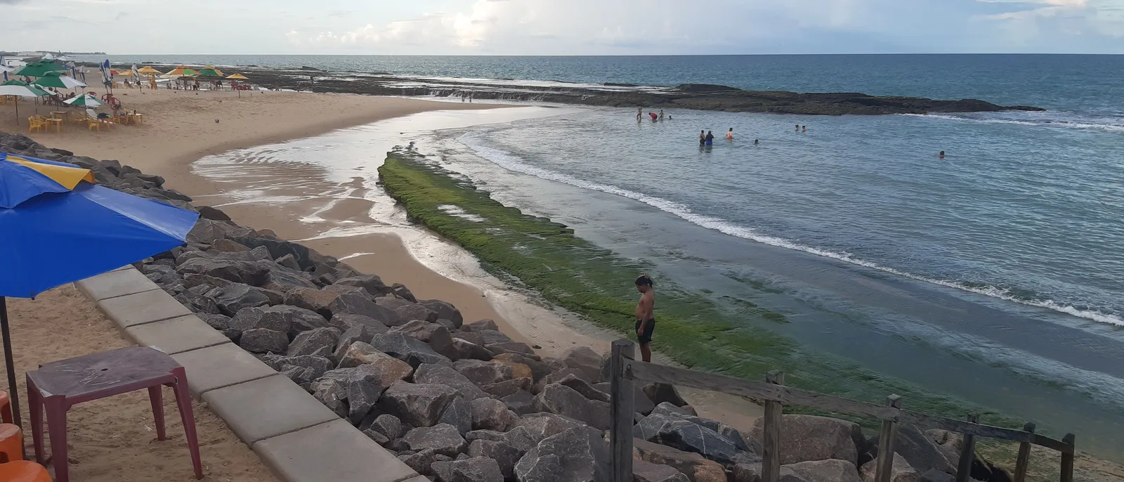 A rock sea wall in the foreground at Praia do Meio in Natal, with a strip of sand curving away past a few bathers and a dark ledge of reef lying offshore