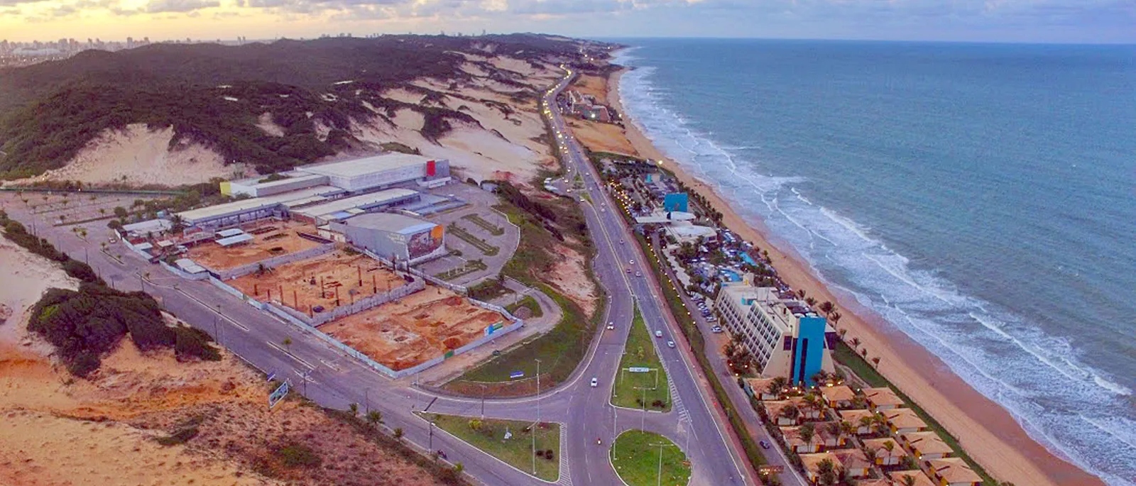 Aerial view along the Via Costeira in Natal: a coast road with the open sea and its beach on one side and a bank of bare sand and dark scrub on the other, and low buildings and parking spread between them