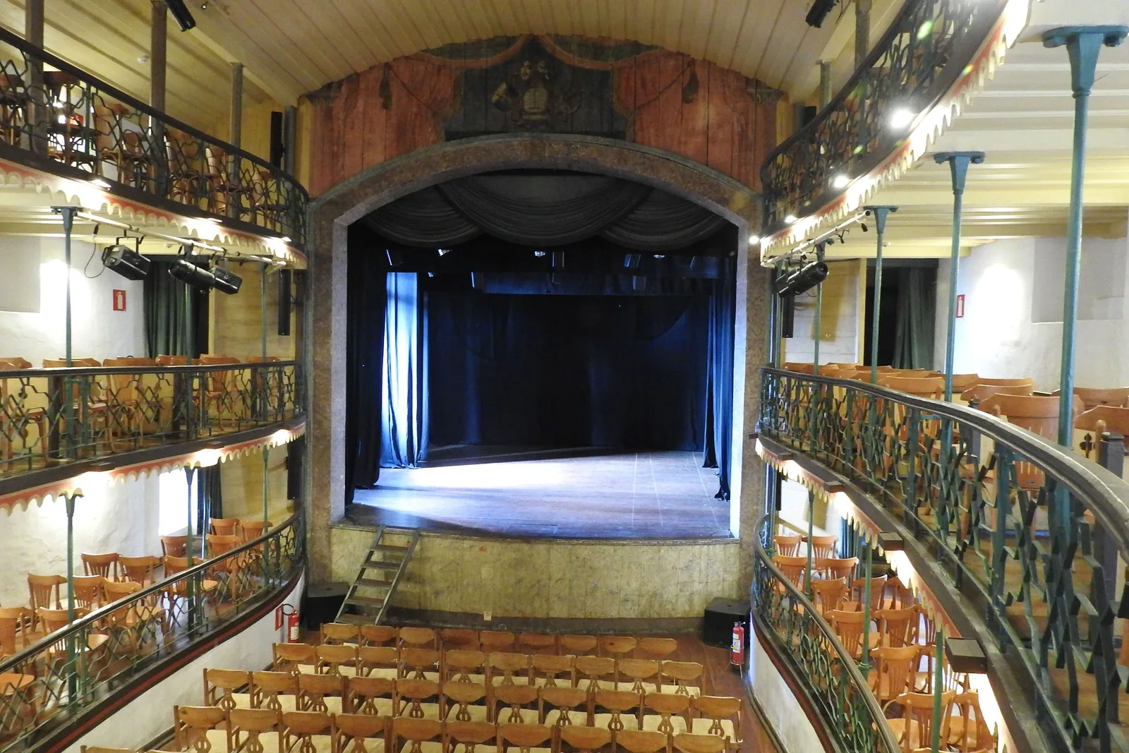 The interior of the Casa da Ópera in Ouro Preto seen from a box: rows of wooden chairs on the raked floor, curved balconies with iron railings running around the walls, and the empty lit stage at the far end