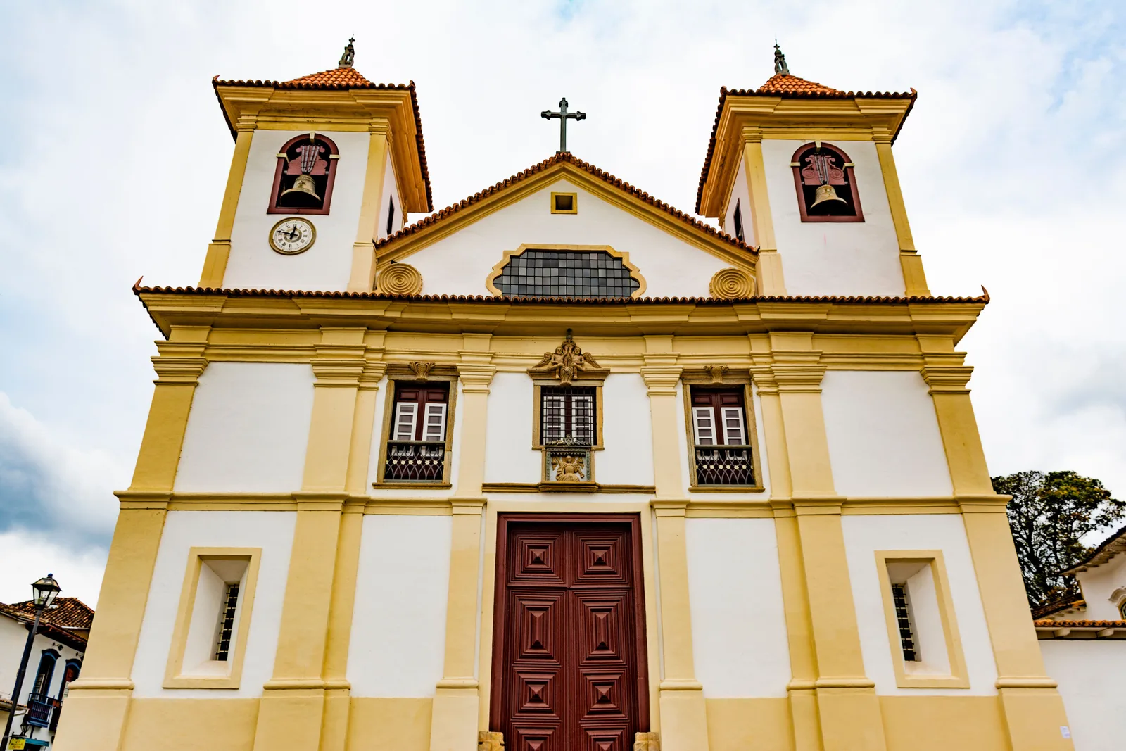 The yellow-and-white facade of Mariana’s Catedral Basílica da Sé in strong afternoon sun, two low bell towers either side of a central pediment and a dark wooden door at the top of the steps