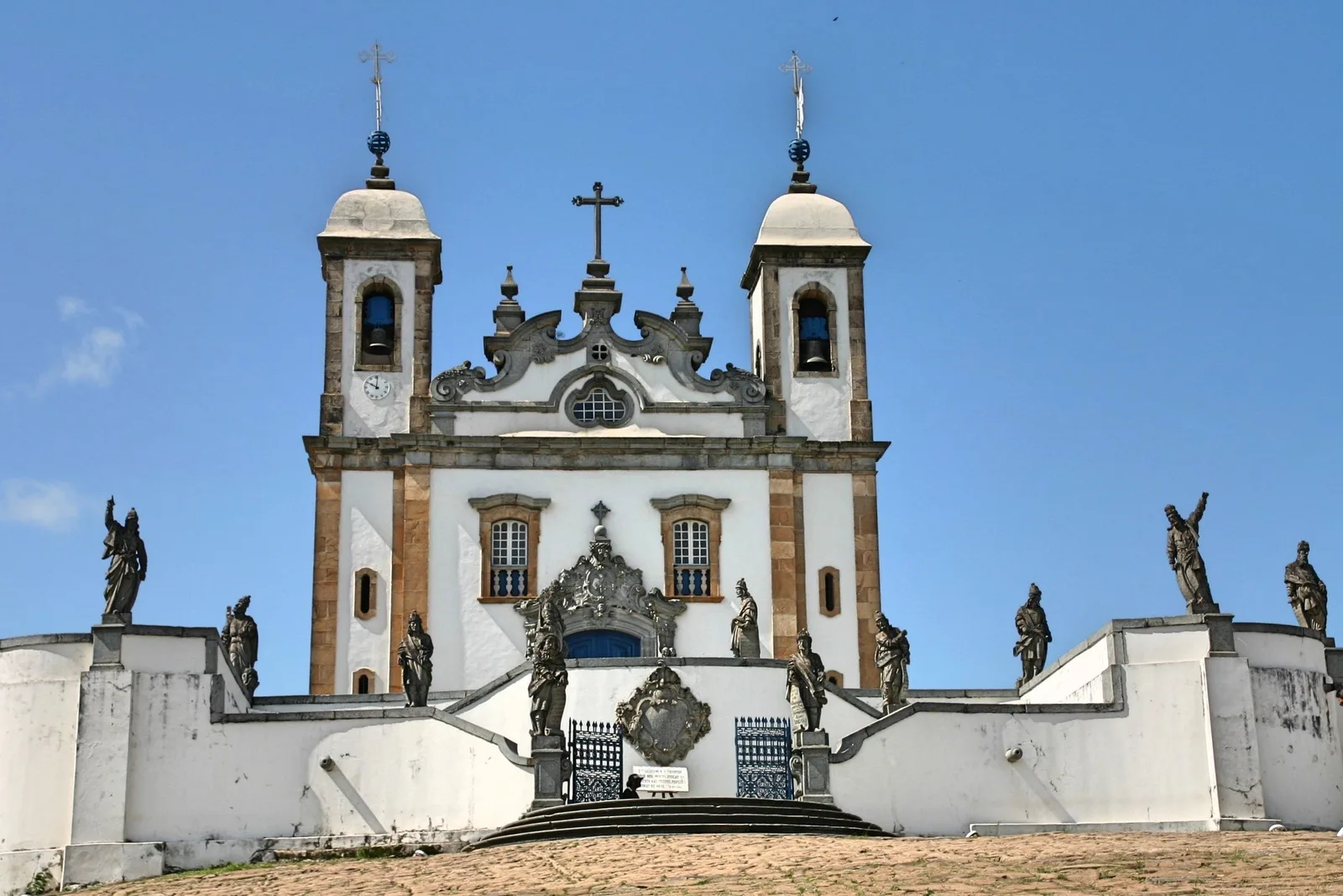 The Santuário do Bom Jesus de Matosinhos at Congonhas straight on in bright sun: a white church with two bell towers behind a low curving wall, soapstone prophets ranged along the balustrade and around the stone stairway in front of it
