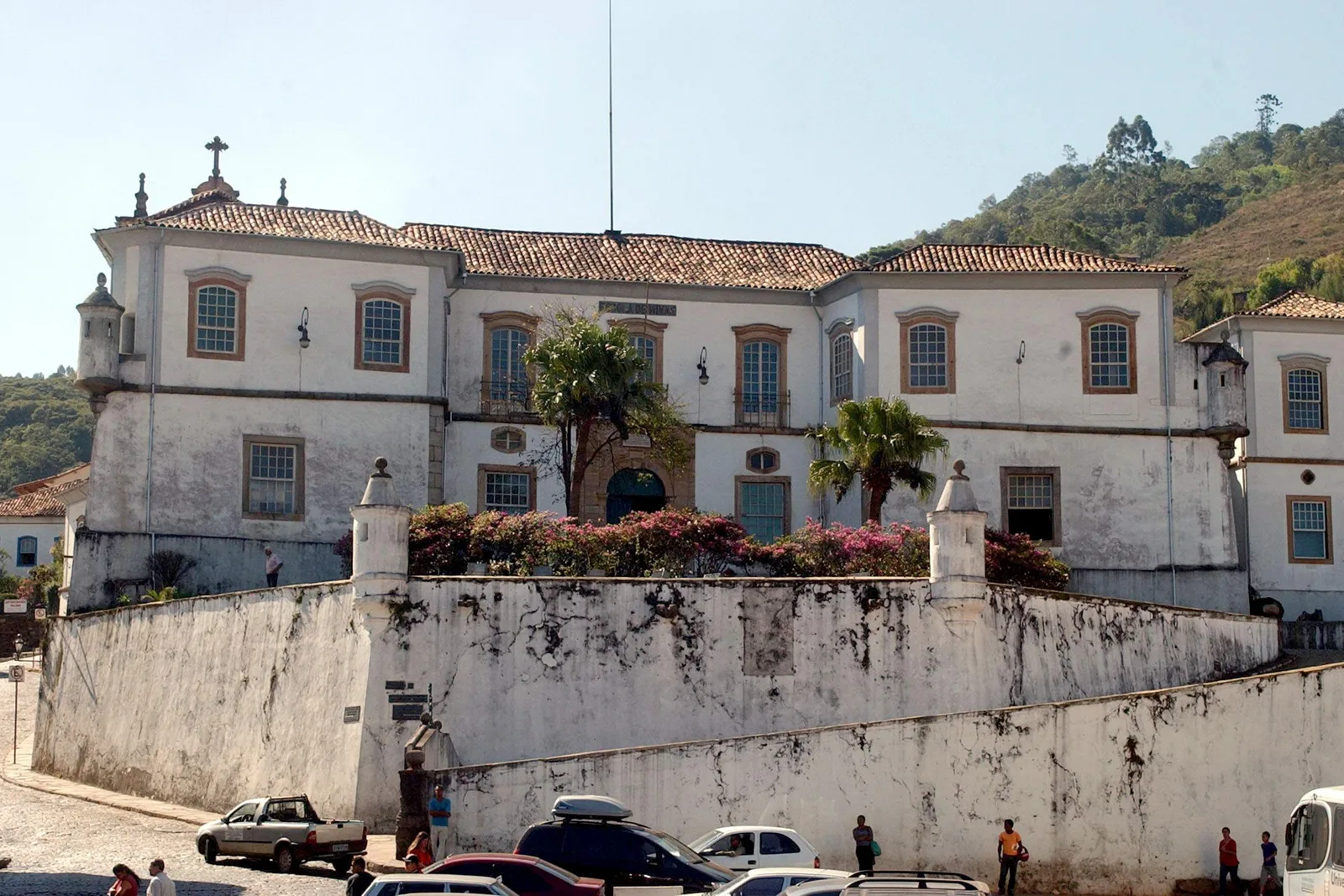 The long white two-story facade of the old Palácio dos Governadores in Ouro Preto, now the Escola de Minas, standing behind a high curved retaining wall with cars parked along the street below it